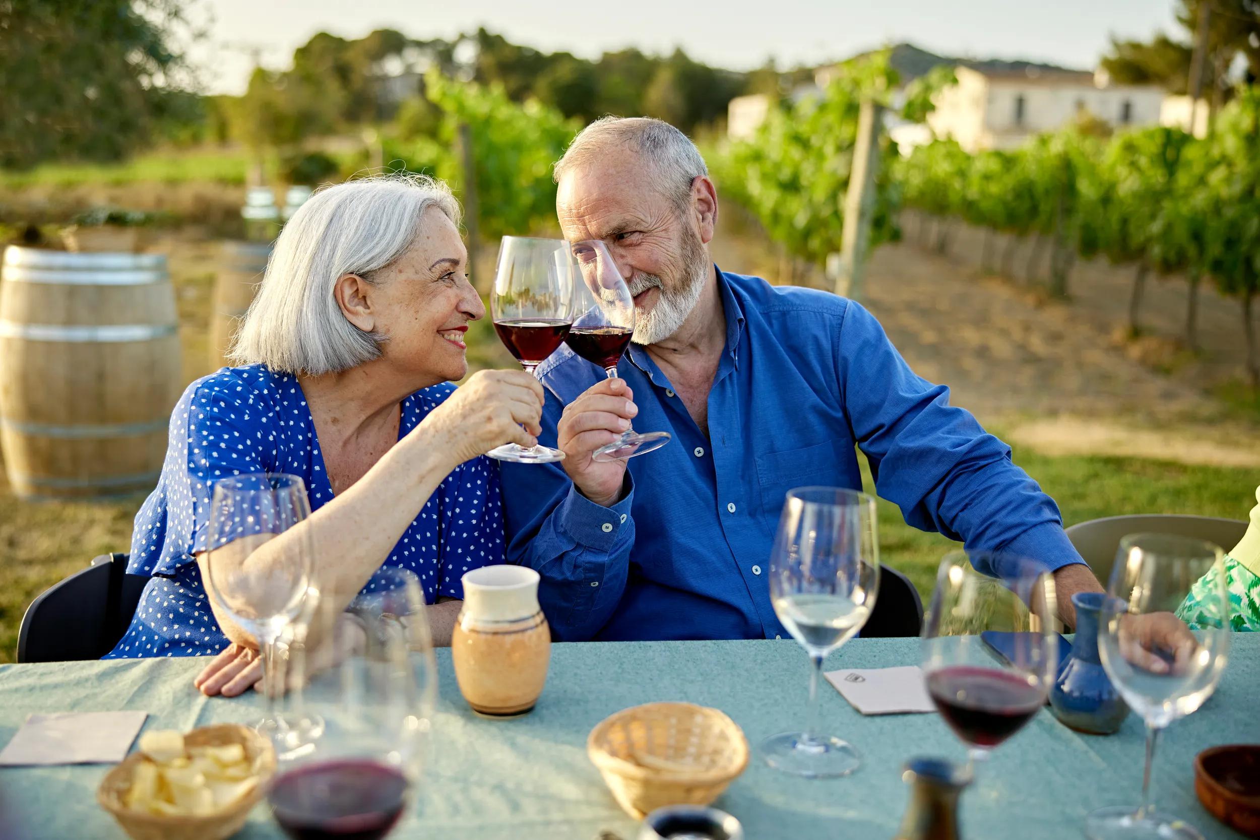 Waist-up view of vacationers sitting at table, holding glasses of red wine, and smiling face to face with winery grounds in background.