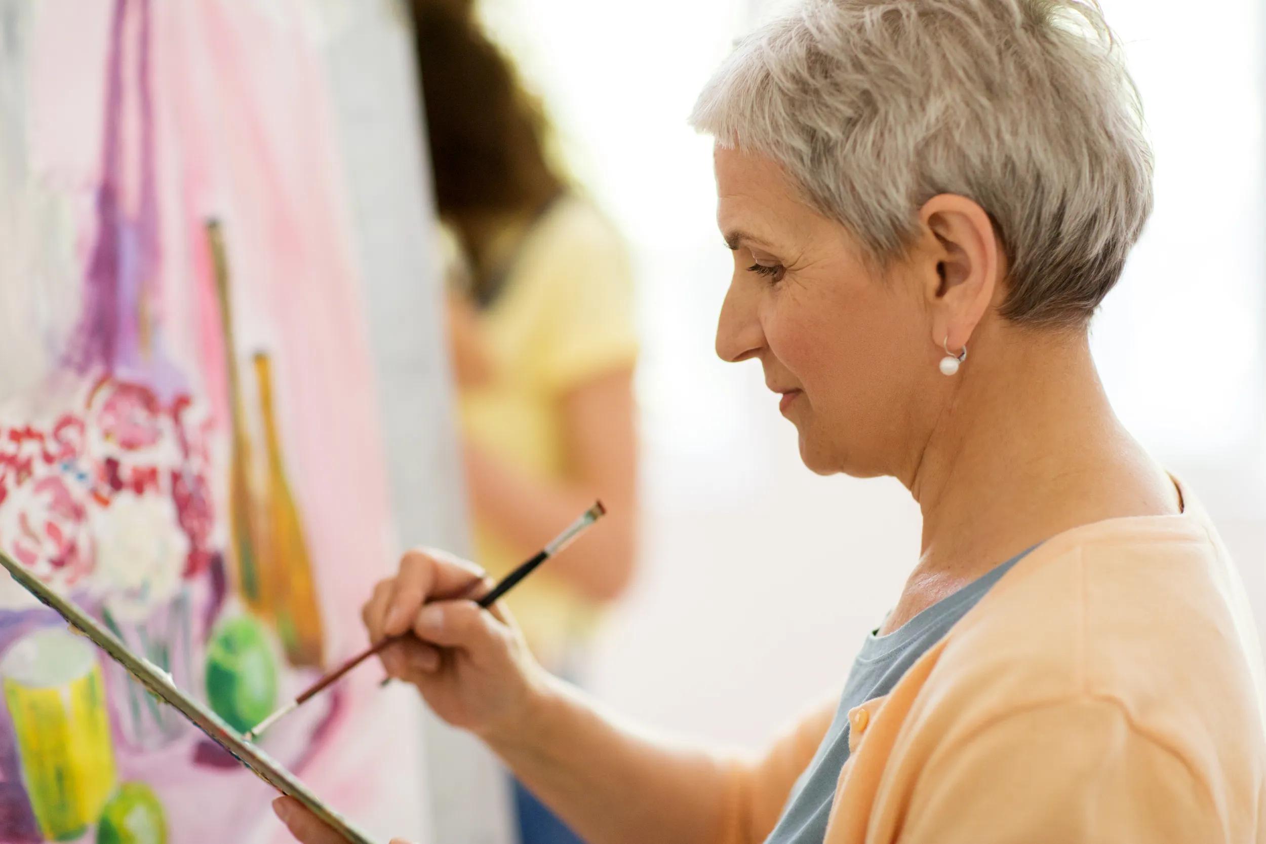 art school, creativity and people concept - happy senior woman artist with easel, paint brush and palette painting at studio