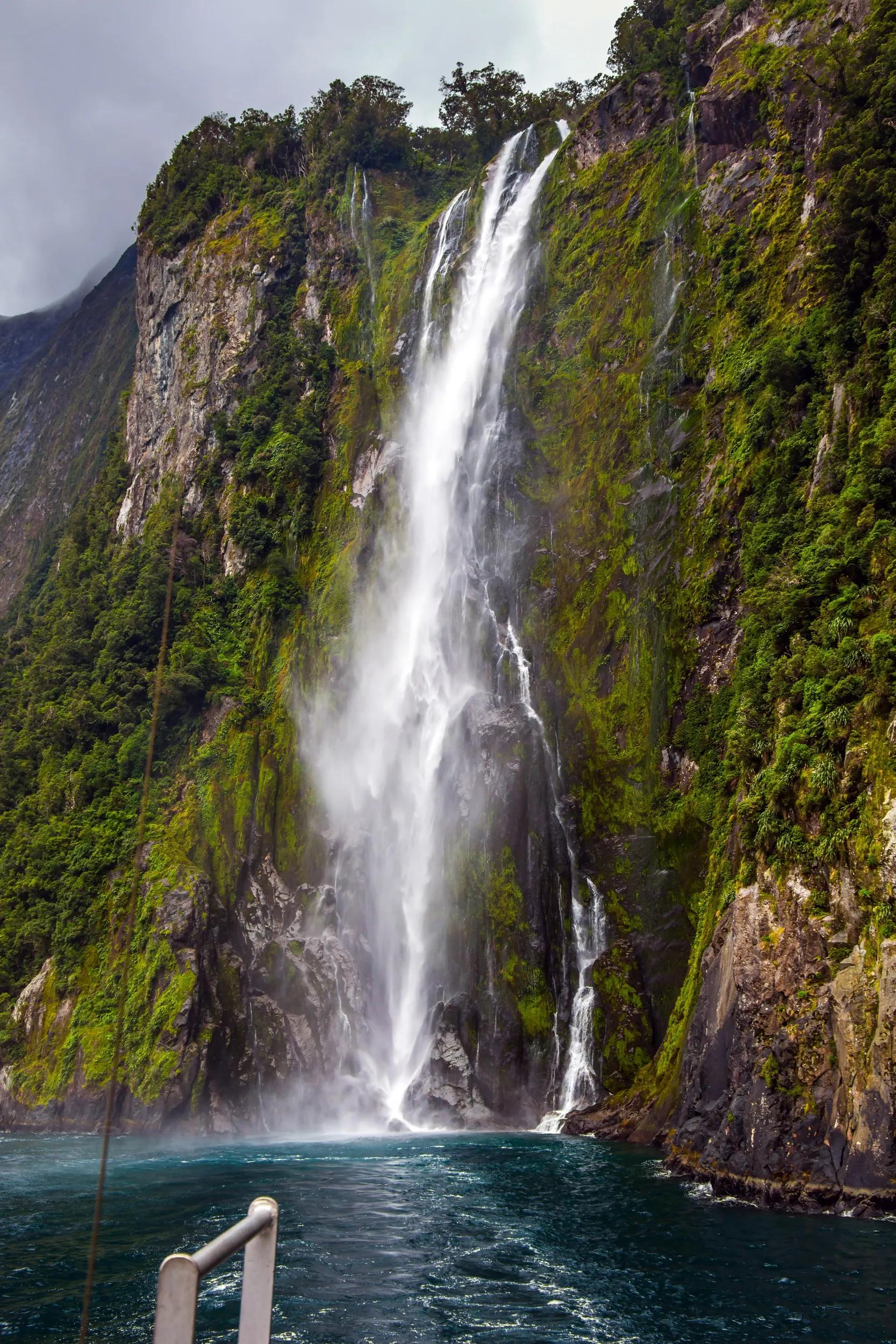 Magnificent waterfall crashes down from the cliff. Tourist cruise aboard the Milford Sound fjord. Magical journey to New Zealand. Concept of exotic, active and photographic tourism