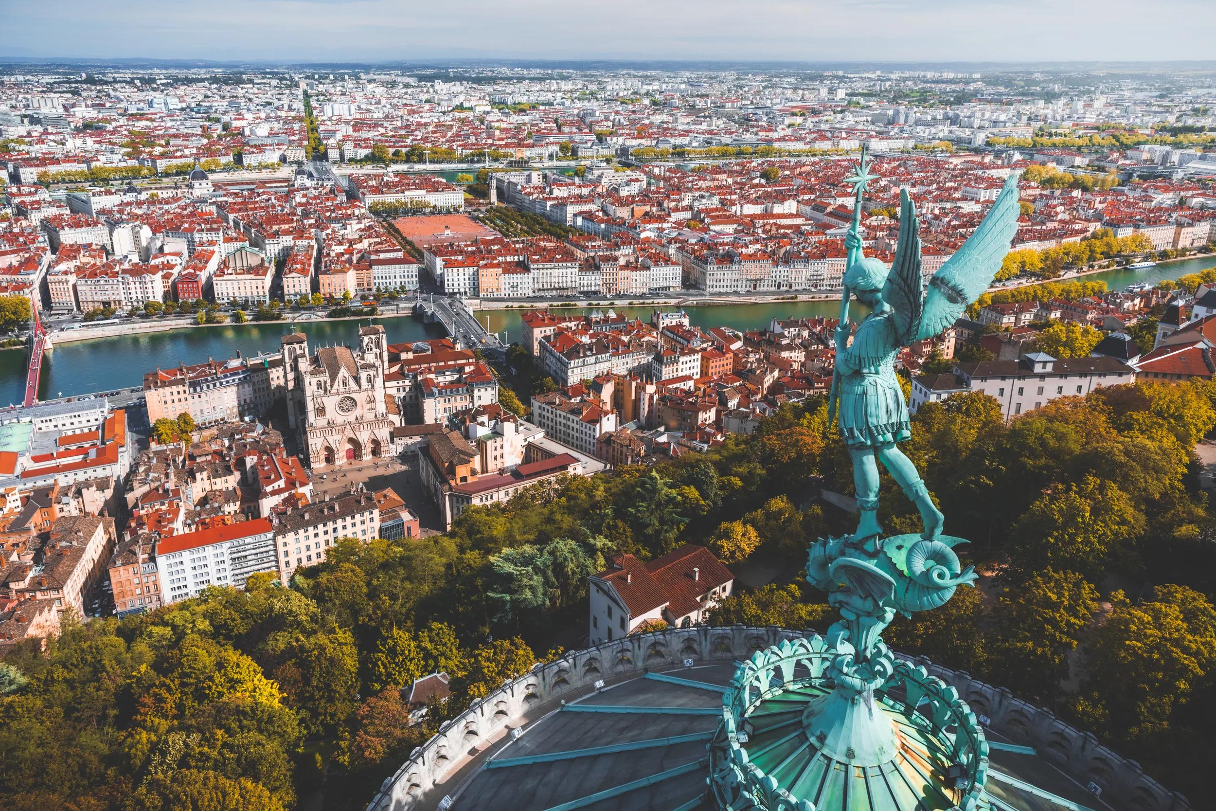 Awesome aerial view on Lyon cityscape, with Saint Jean-Baptiste Cathedral, both Saone and Rhone river, with Bonaparte bridge, famous place Bellecour and typical red roofs from apartment residential buildings. High angle view of Lyon viewed from the roofs of Basilica Notre Dame de Fourviere with verdigris Archangel Michael statue overlooking the city. The Basilica was built between 1872 and 1884, dedicated to the Virgin Mary. This image was taken during a sunny summer day in Lyon city in Rhone department, Auvergne-Rhone-Alpes region in France (Europe), outside Basilica Notre Dame de Fourviere, on Fourviere hill.