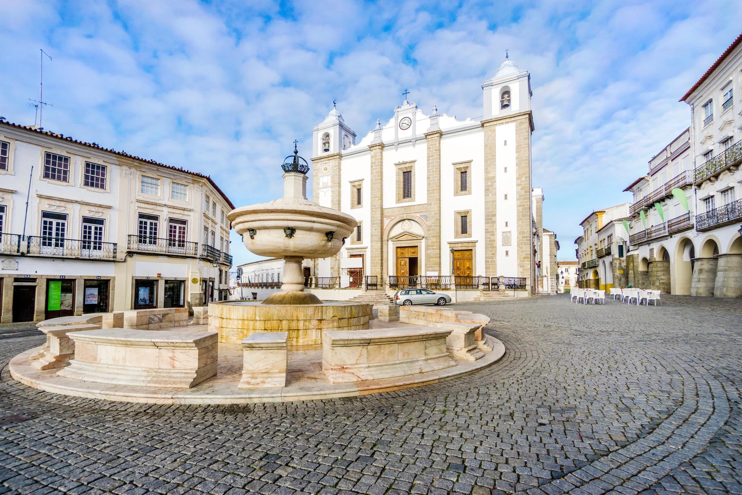 Low-angle shot of a fountain surrounded by traditional residential building and with Saint Anton's Church in the background, Giraldo Square, Evora, Alentejo, Portugal