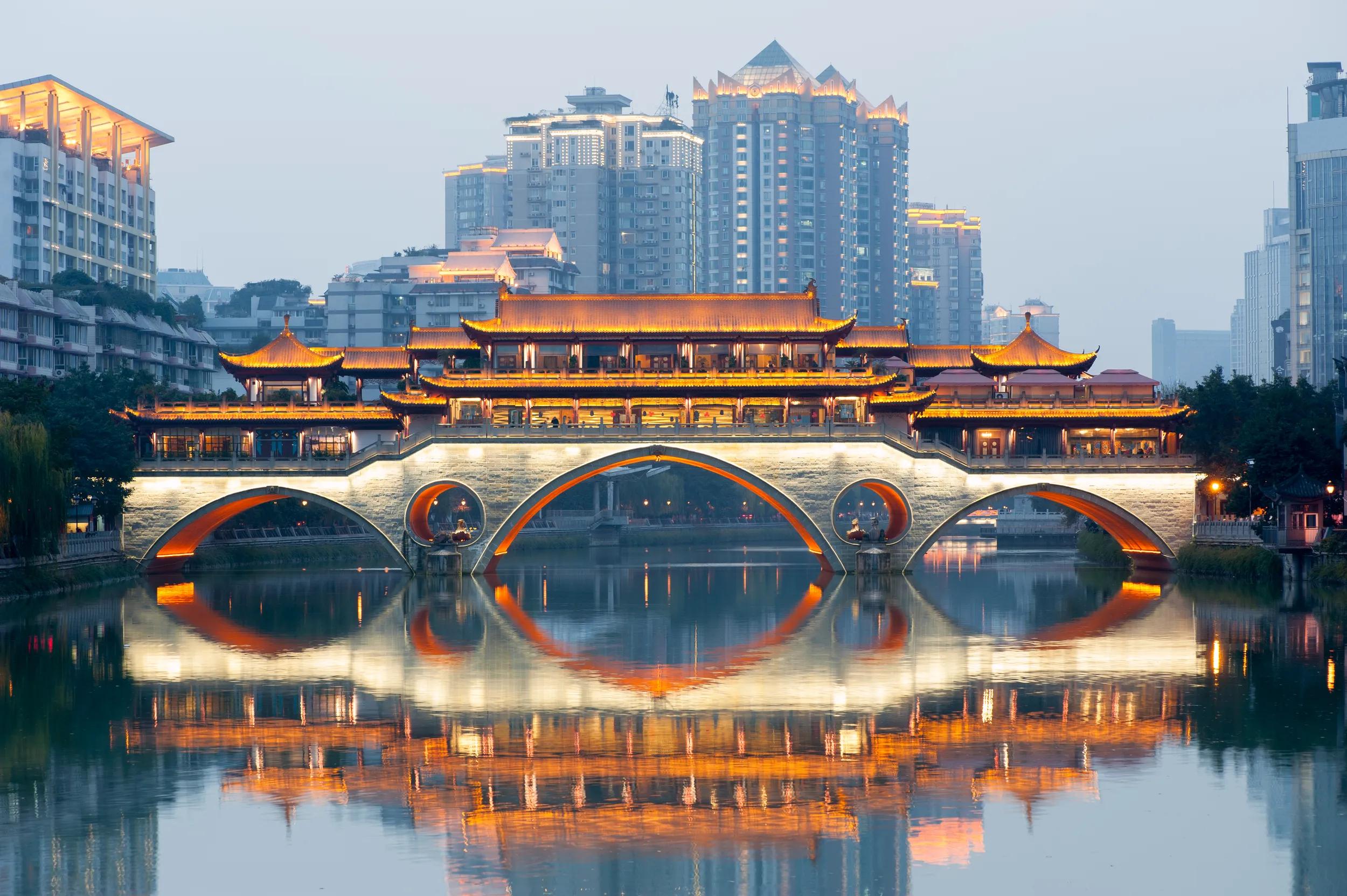 Anshun bridge with lights reflecting in the river at dawn with buildings in blue haze in the background, Chengdu, China.