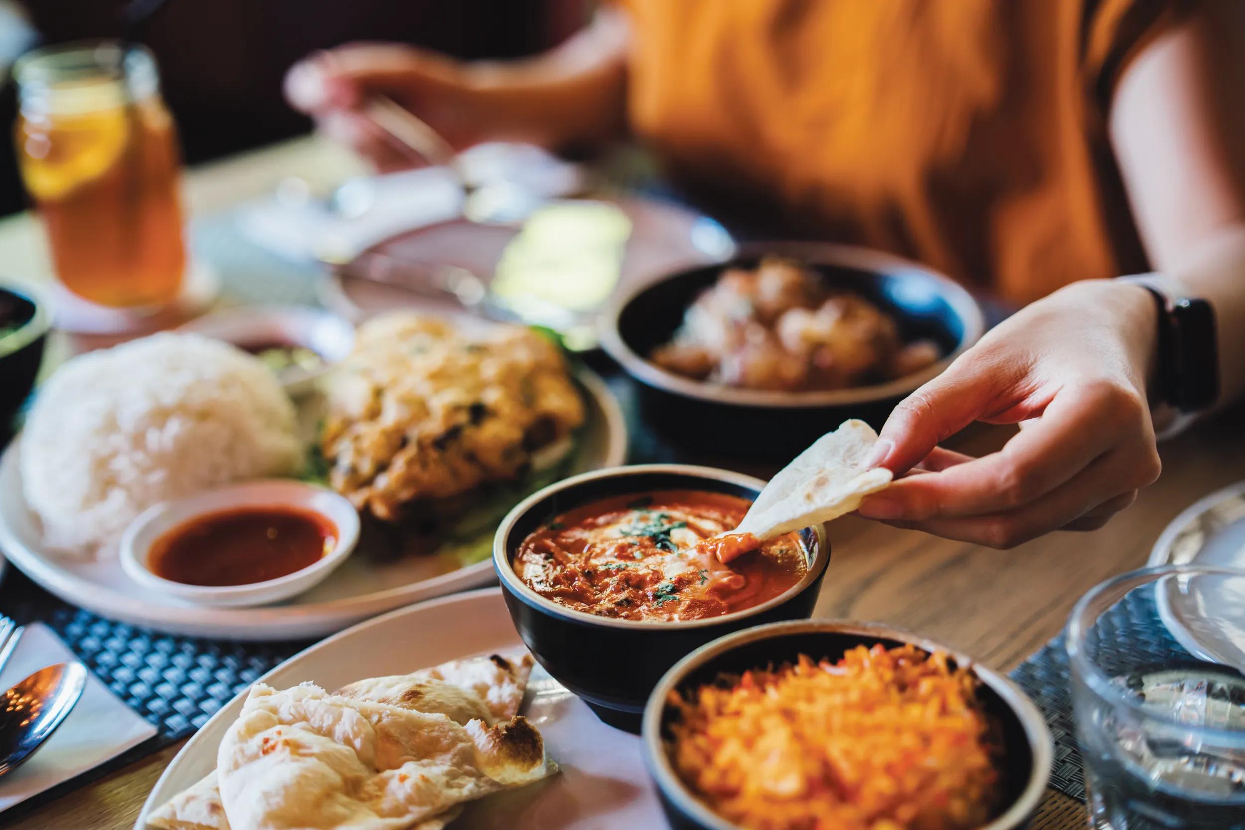 Young Asian woman tasting authentic classic Indian food with assorted dishes freshly served on the table in restaurant. Close-up of her hand dipping naan bread on the butter chicken. Tourist exploring local cuisines when travel. People, food and lifestyle