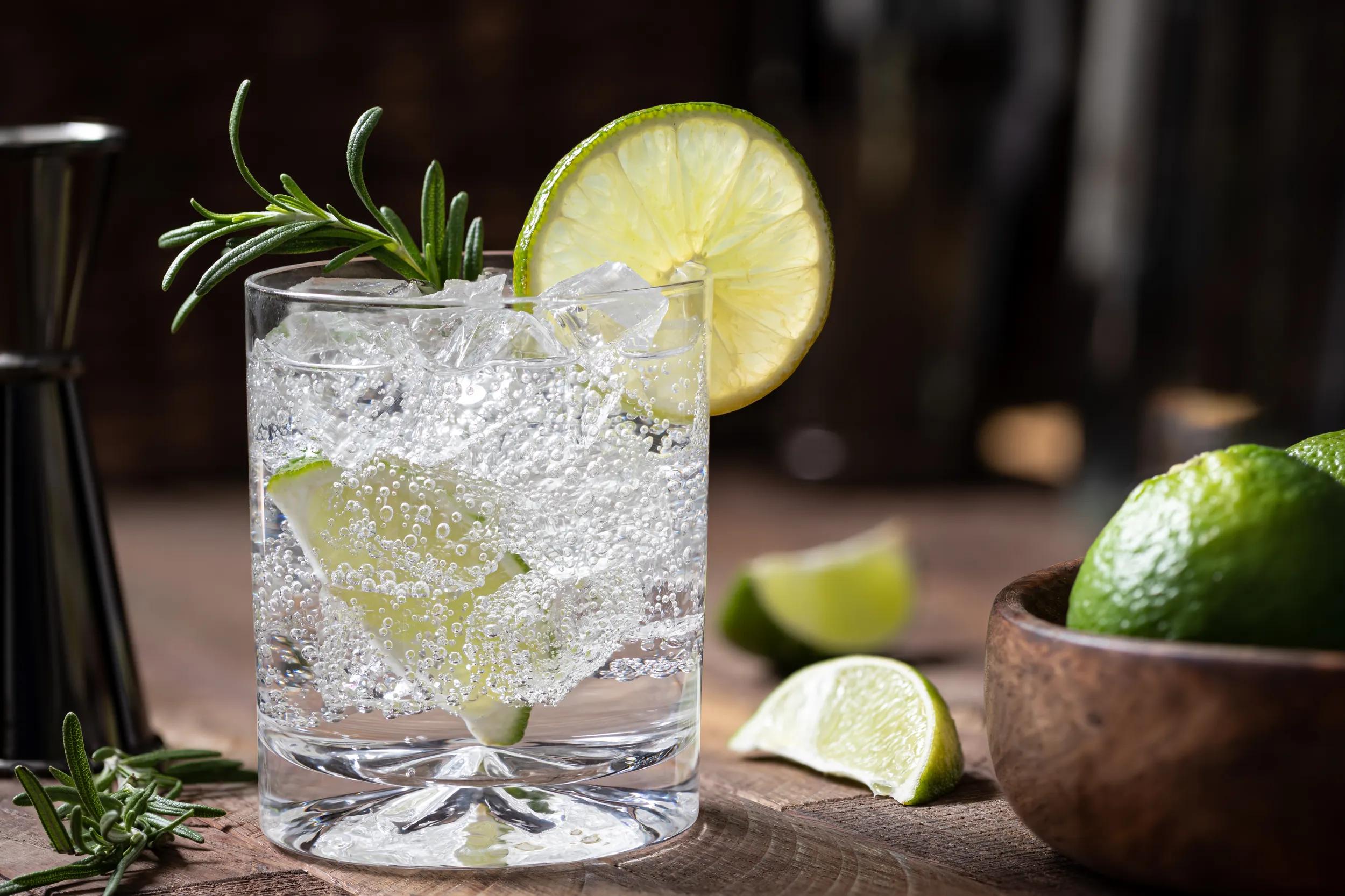 Gin and tonic cocktail with lime slices, rosemary and ice on a rustic wooden table