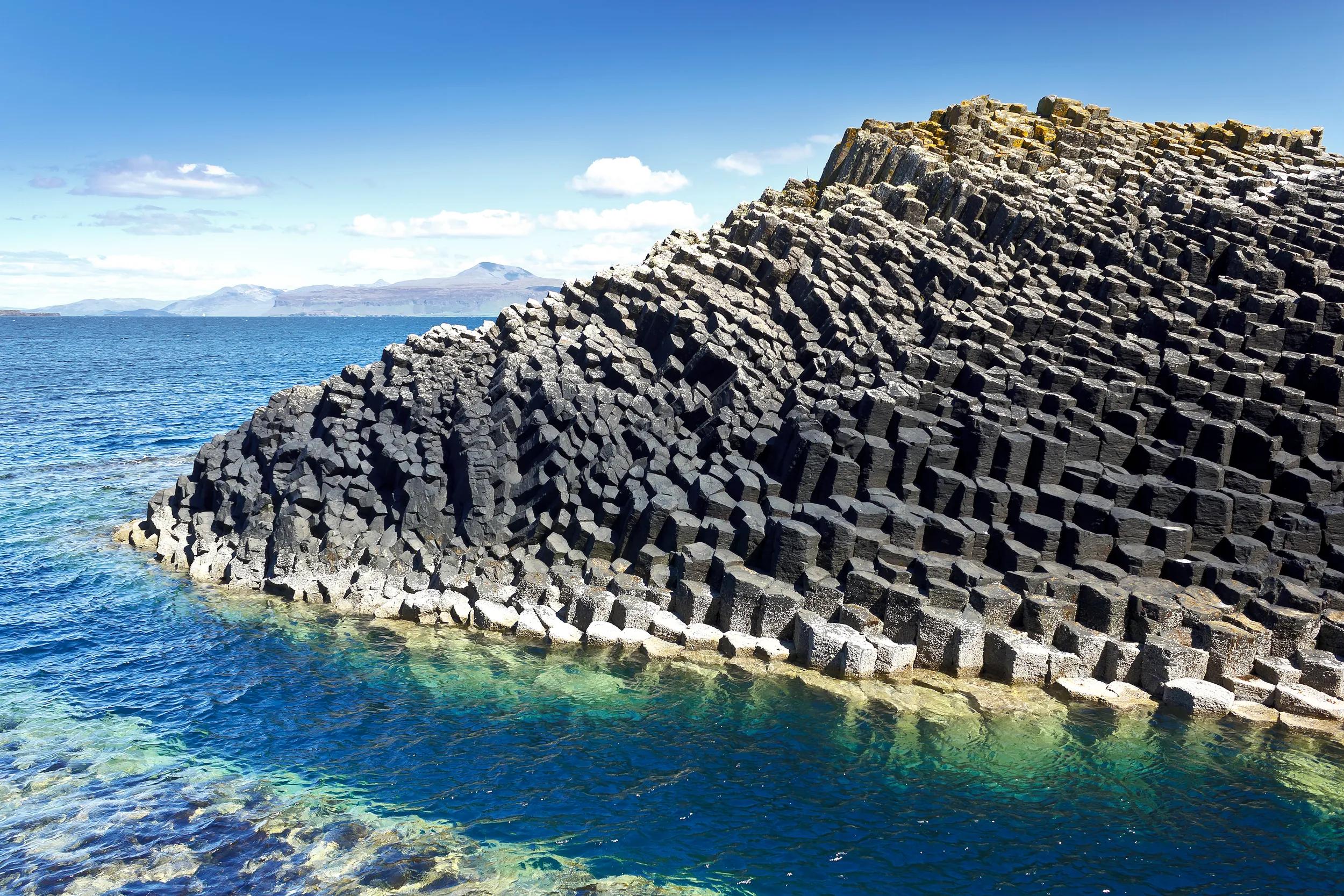 Isle of Staffa, Scotland: dark hexagonal basalt columns in the turquoise waters of the Inner Hebrides against a blue summer sky.