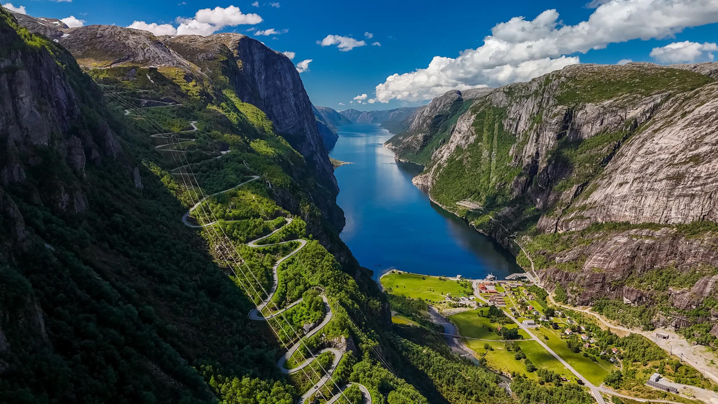 An aerial view of a winding road snaking through the lush green mountainsides overlooking a tranquil Norwegian fjord on a sunny day. Kjerag, Lysebotn, Lysefjorden, Norway