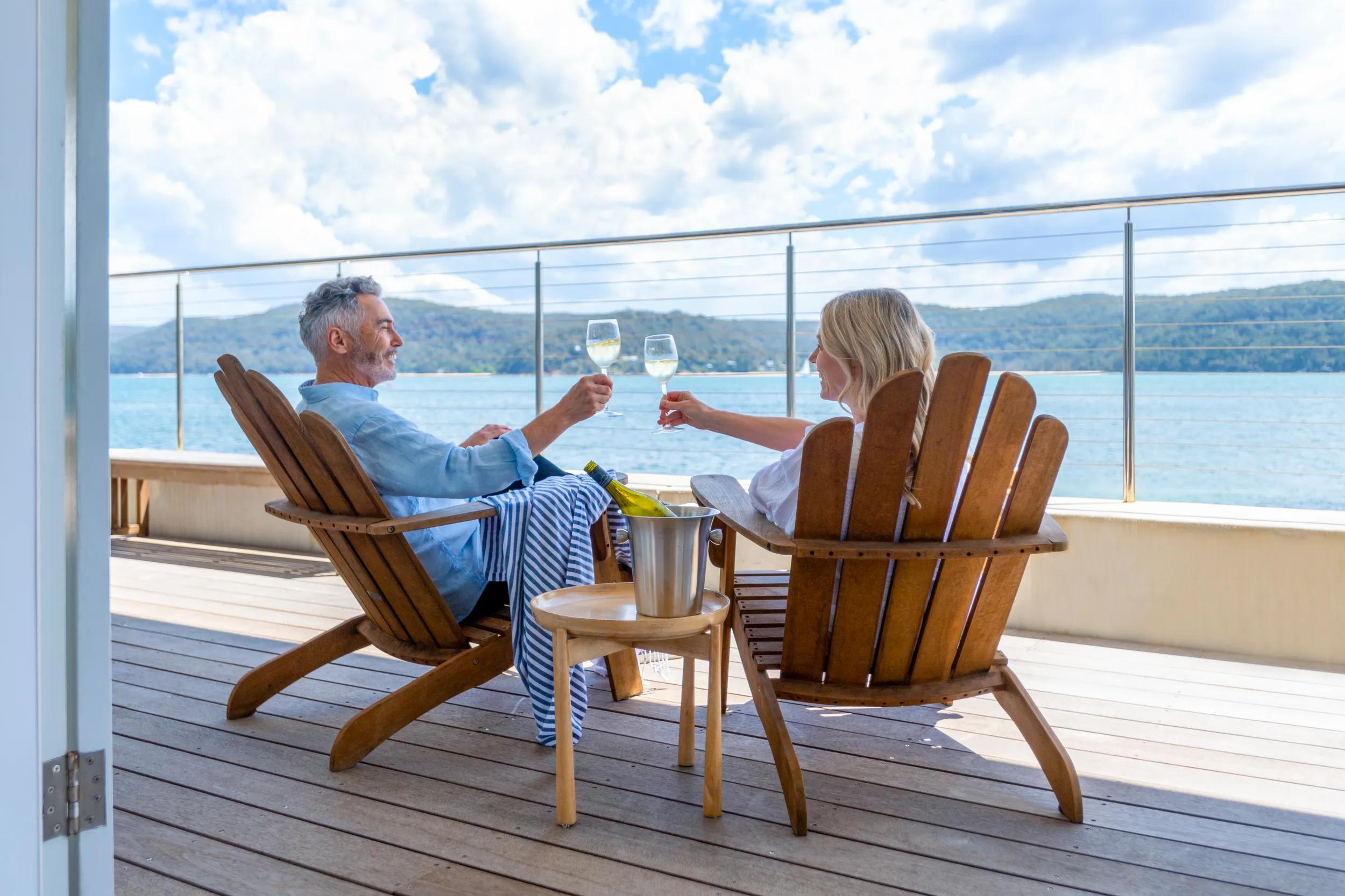 Mature couple drinking wine out on the deck. They are happy and smiling sitting in deck chairs. The sea is in the background