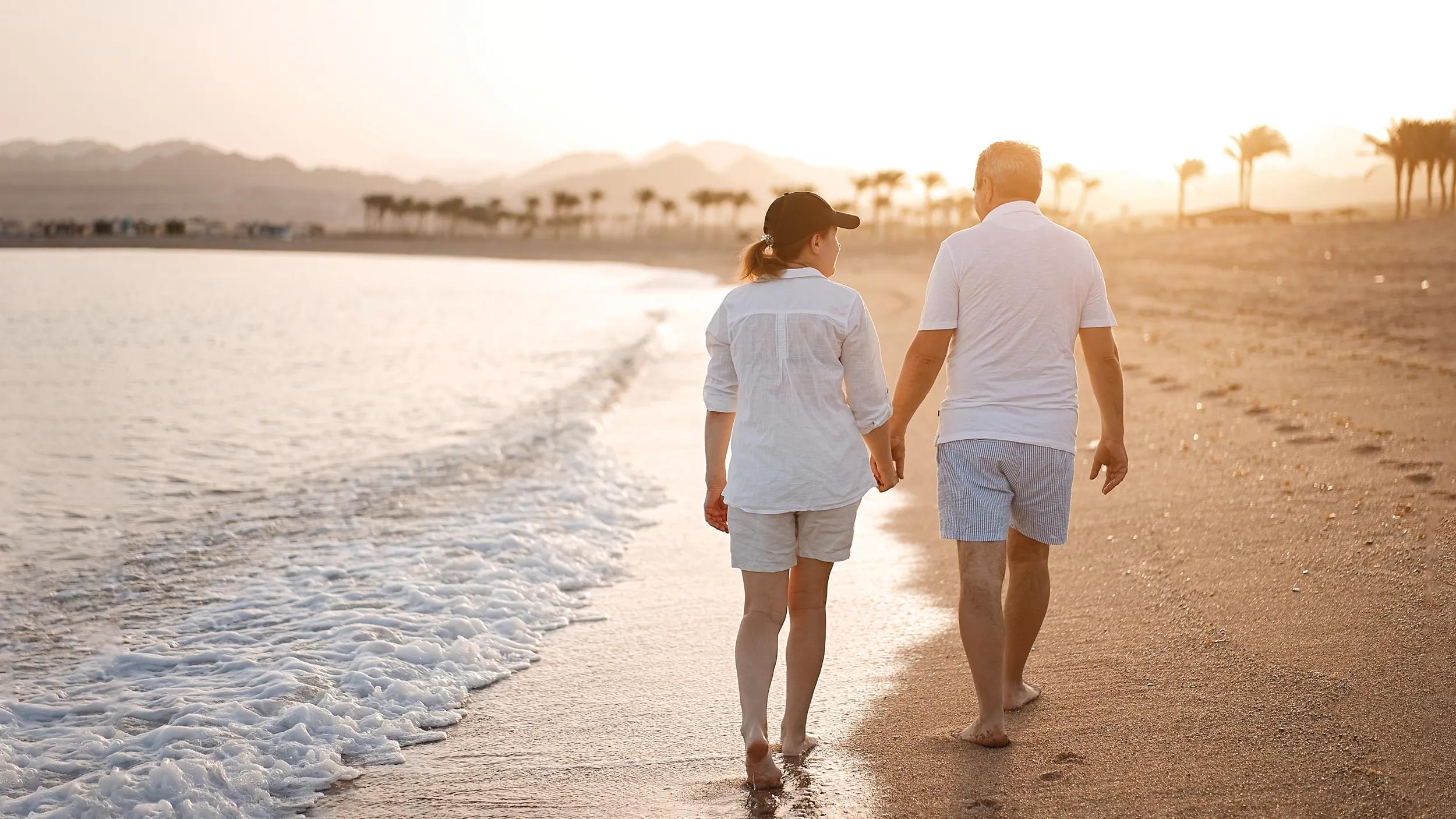 Happy mature senior couple walking and looking at each other on beach during sunset. Aging together and retirement lifestyle concept