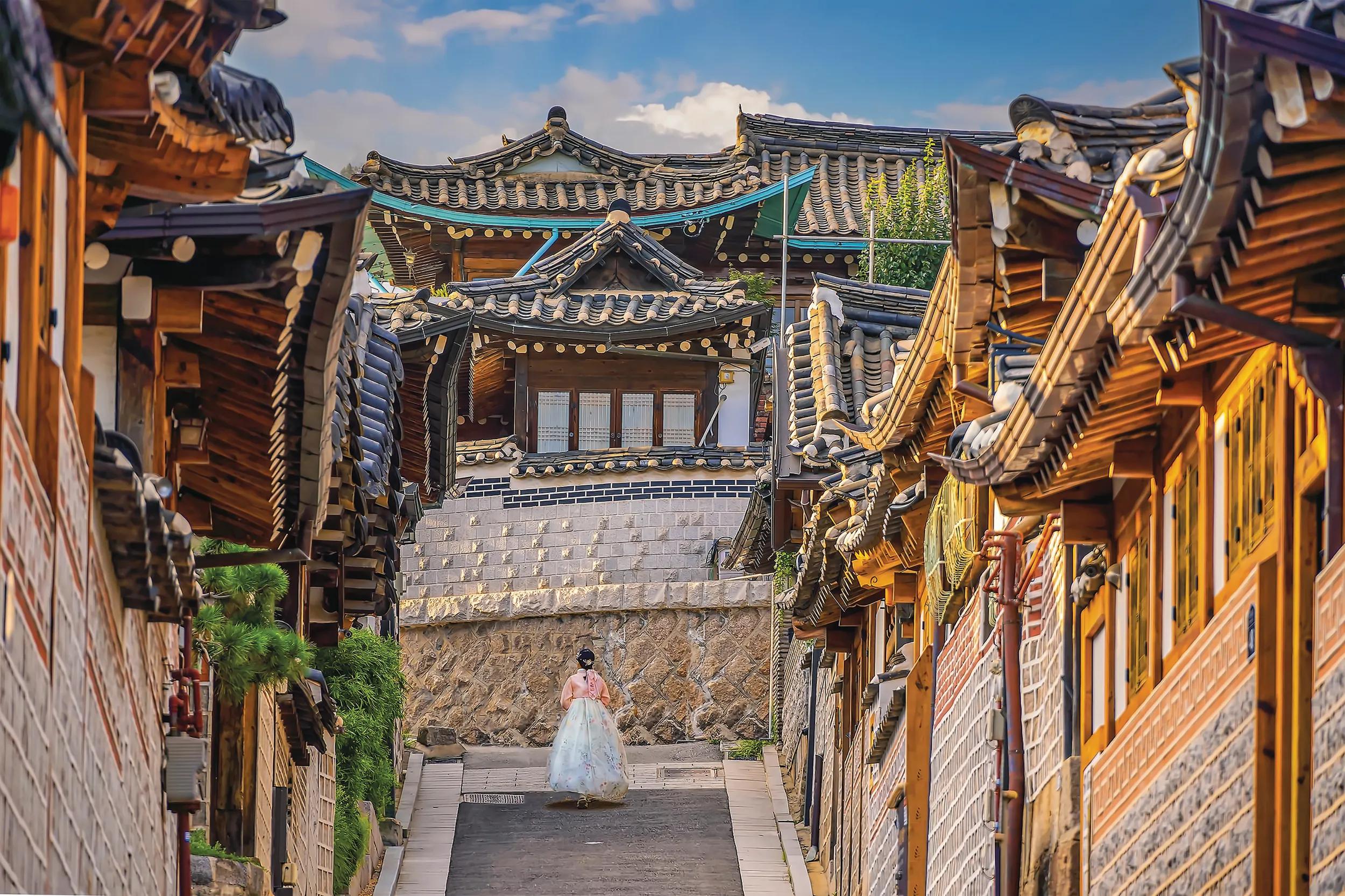 Girl wearing hanbok walking through Bukchon Hanok Village with Seoul city skyline, cityscape of South Korea at sunset