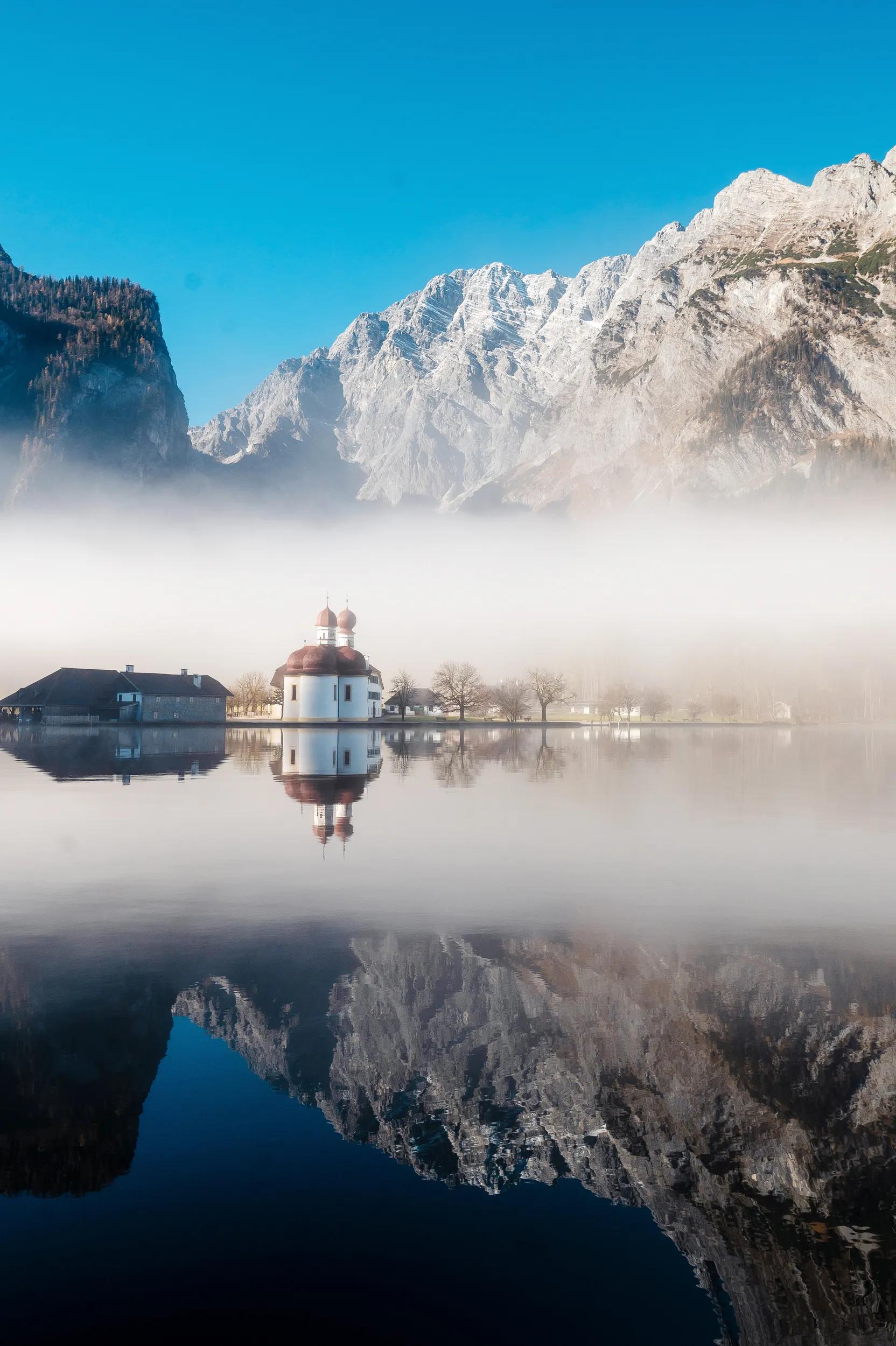 Strong morning fog over church St. Bartholomew on Konigsee lake during winter time. Beautiful rocky alps on background with clear reflection on water.