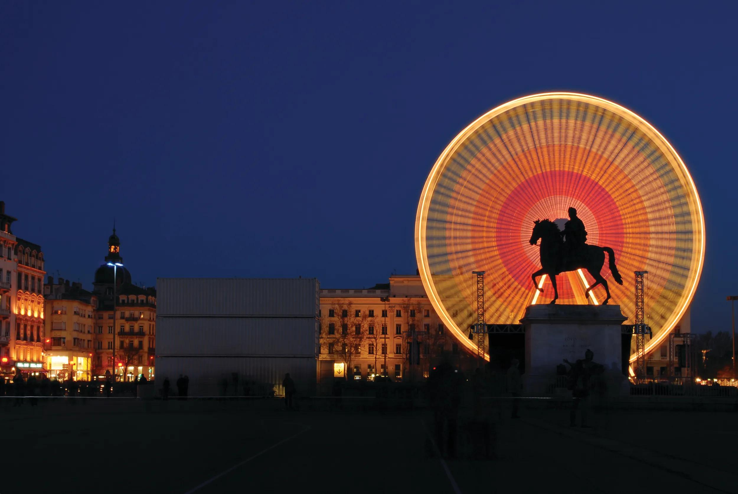 La Place Bellecour is a large square in the centre of Lyon, France. Equestrian statue of Louis XIV in silhouette. Long exposure created blurred Ferris wheel.