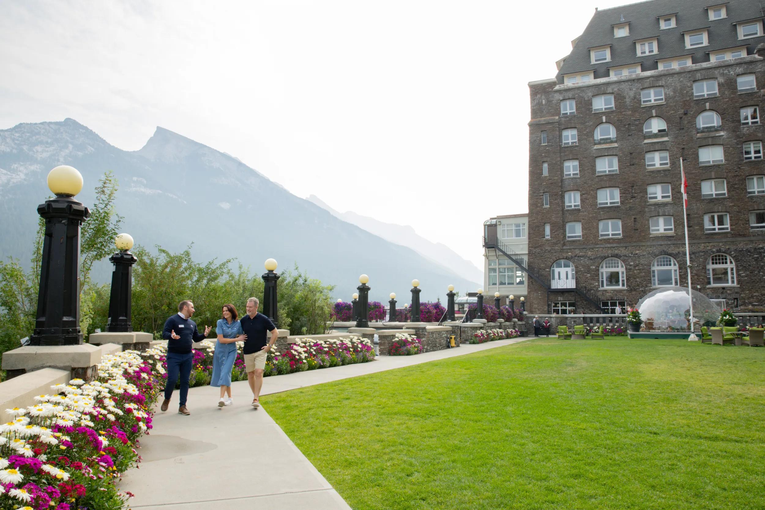Couple with APT Tour Director Zac Watts on the grounds of the Fairmont Banff Springs Hotel with views over the Canadian Rocky Mountains.