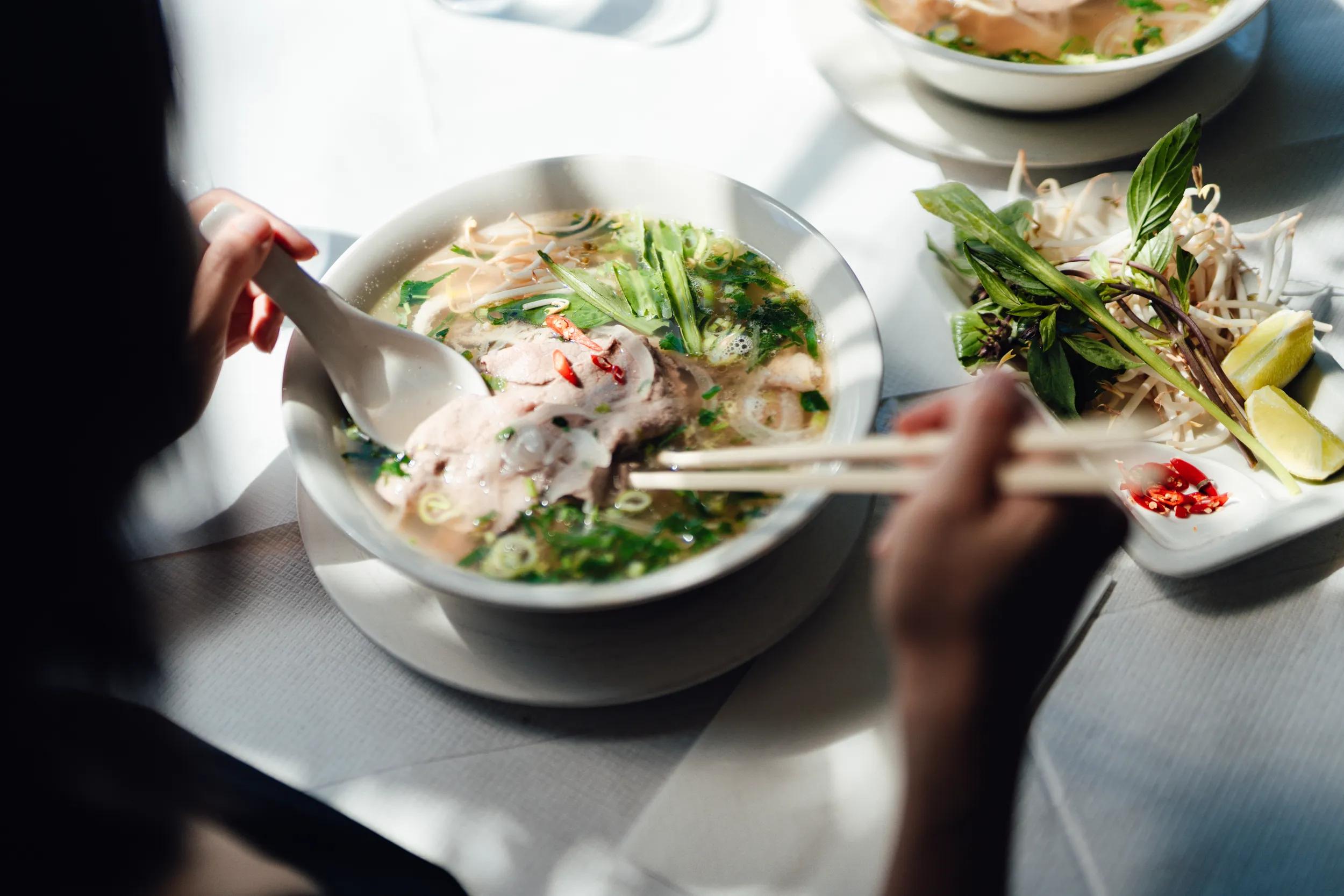 Over the shoulder view of young woman eating traditional Vietnamese pho soup with chopsticks in the restaurant.