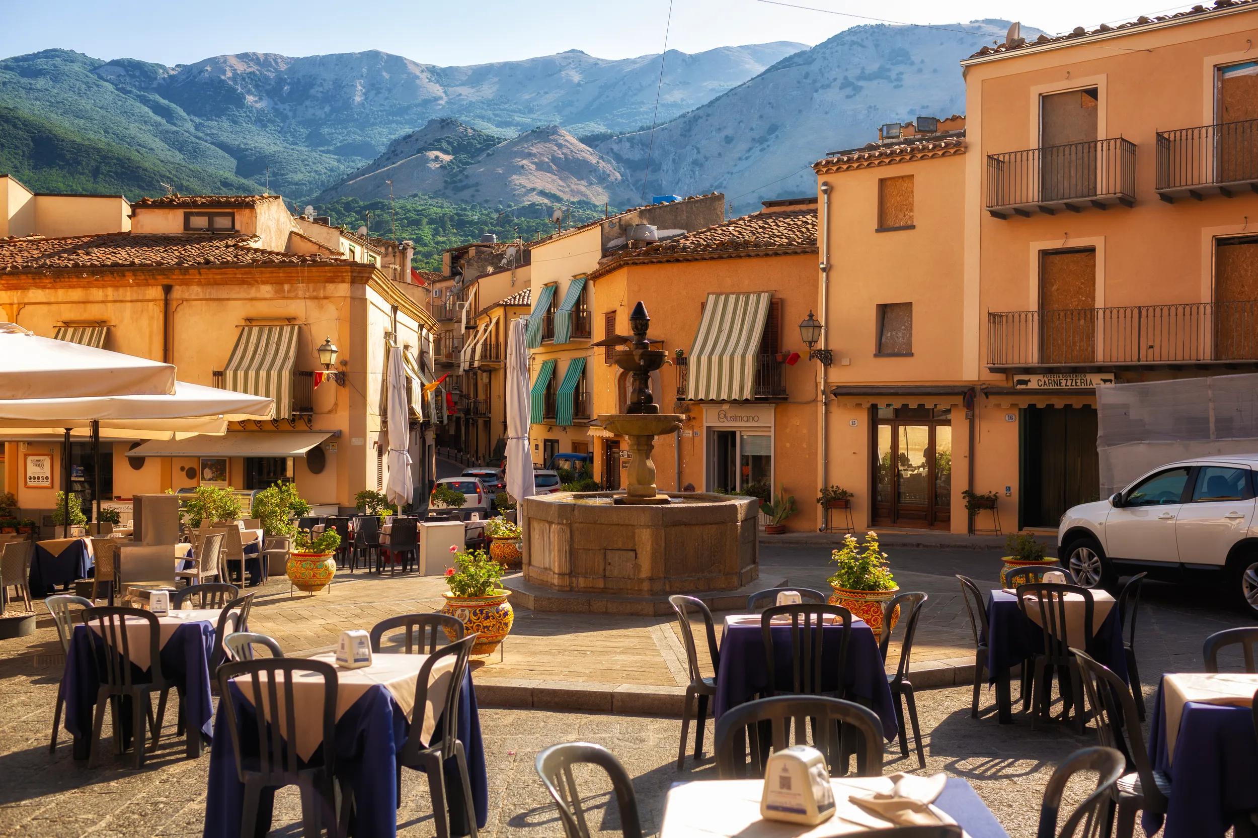 Castelbuono, ITALY - August, 4, 2023. Fountain and cafes with tables in the small town square - piazza of Castelbuono, Sicily. Palermo Province, Madonie mountains on background.