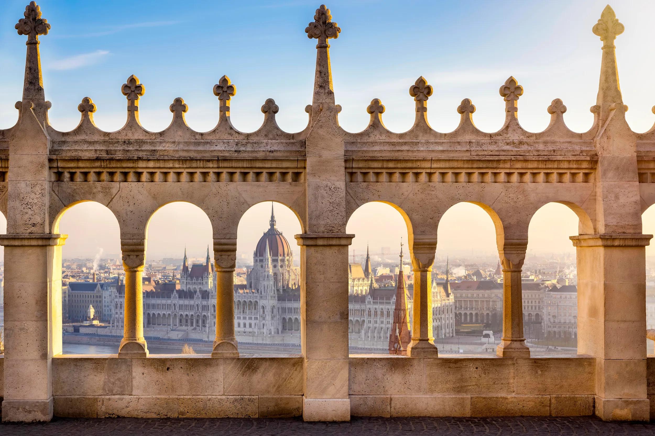 View through the columns of the Fishermen Bastion to the Parliament building at the Danube River in Budapest, Hungary, during golden sunrise time