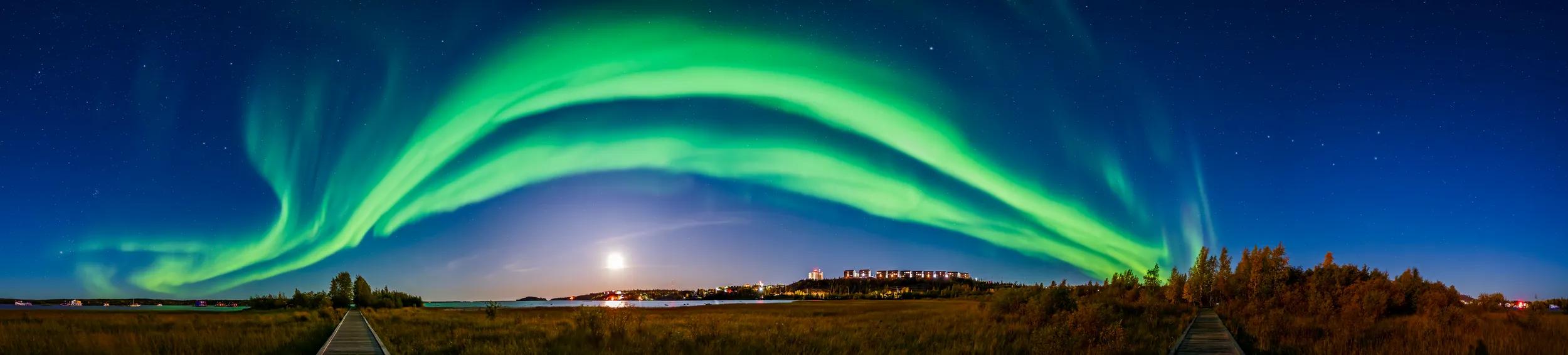 September 10, 2019 - A 240 degree panorama of the northern lights from the boardwalk in the urban Rotary Park in Yellowknife, Northwest Territories, Canada. A waxing gibbous moon is bright to the south and lights the sky and landscape. Despite the moonlight and city lights the aurora still shines brightly. The aurora sweeps across the sky from east to west here, passing high across the south and framing the moon. The Big Dipper is at far right; the Pleiades are rising at far left. Andromeda is at top left.