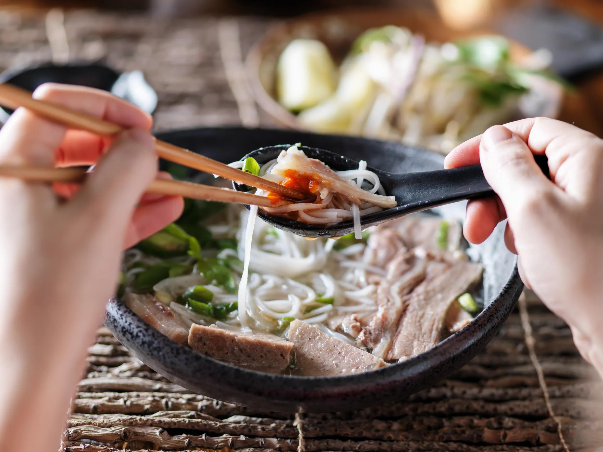 Woman eating pho with sriracha using chopsticks and spoon together.