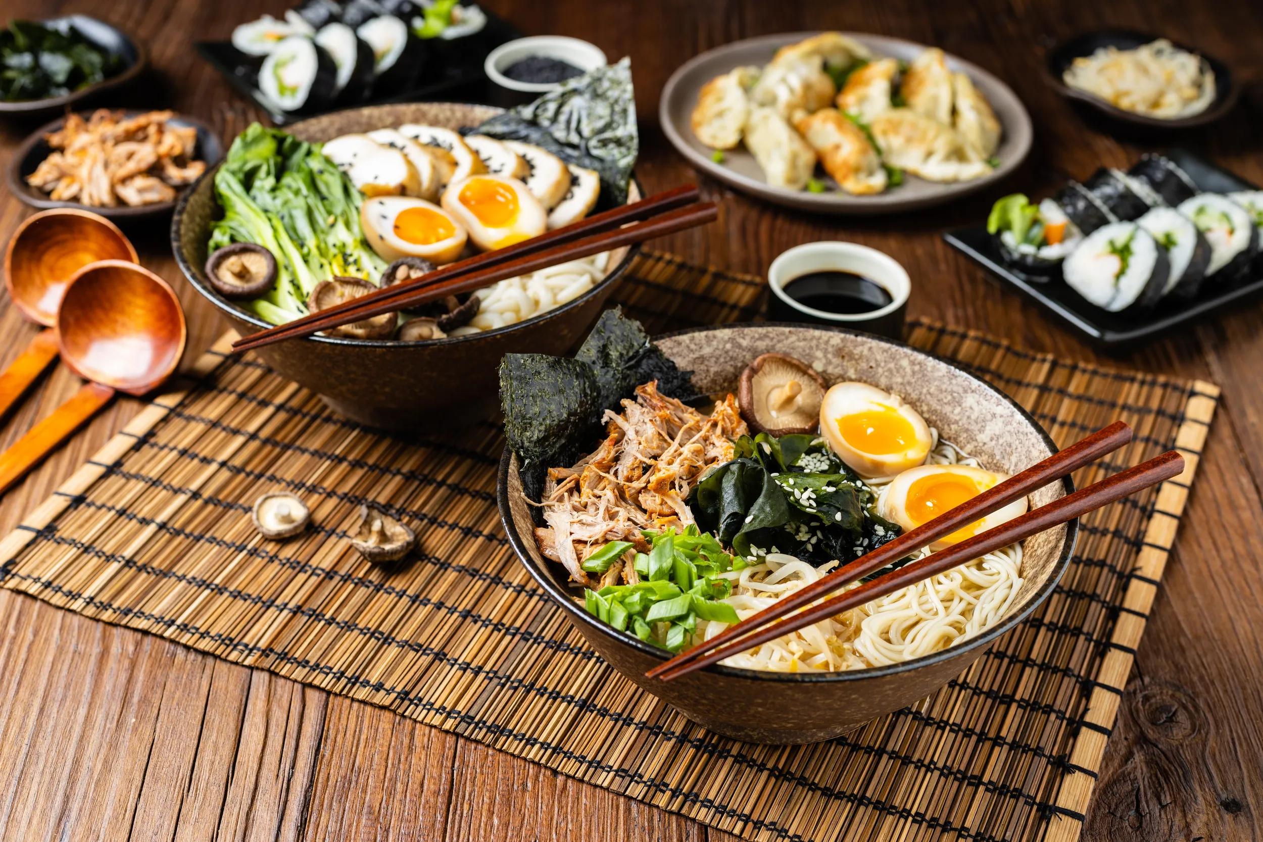 Traditional ramen with jerked pork or chicken.  With udon or ramen noodles. Served in classic bowls. Gyoza dumplings and mushrooms in the background.  Natural wooden background.