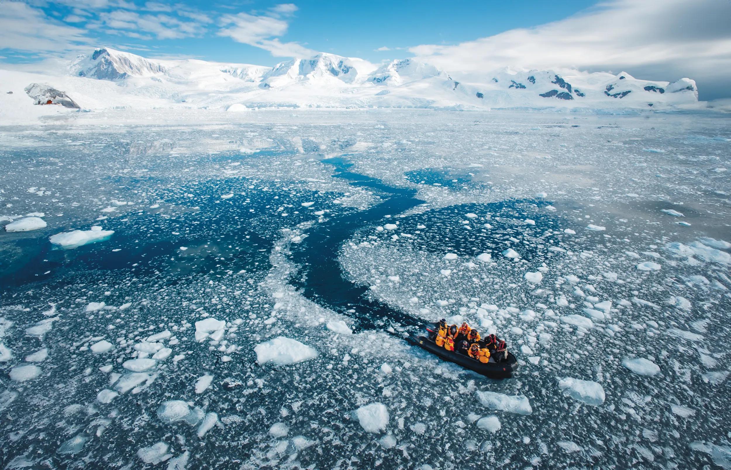 Team of unrecognizable Explorer returning to the Icebreaker Ship from a landing at the Antarctica Peninsula. Cruising with a Zodiac Dinghy through small drift ice and icebergs back to their exploration ship under sunny late morning sky. Antarctic Glaciers and Mountain Range in the background. Antarctica Peninsula, Antarctica