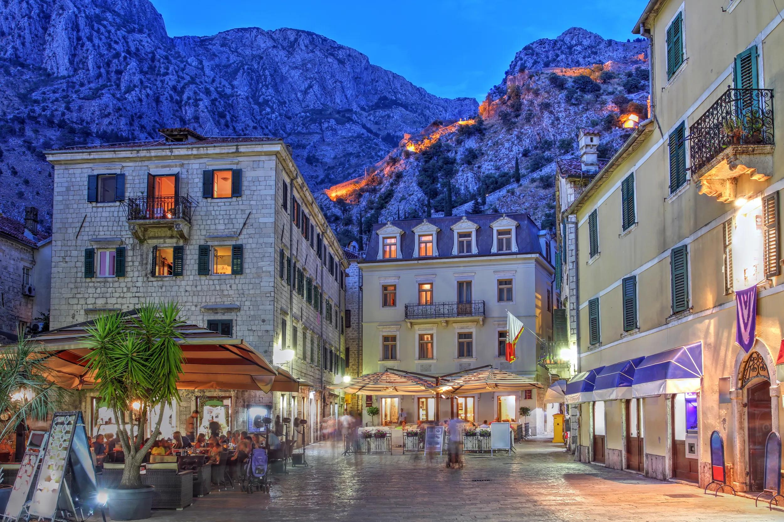 Scene in the medieval town of Kotor, Montenegro at twilight with the ancient city walls illuminated in the background mountains.